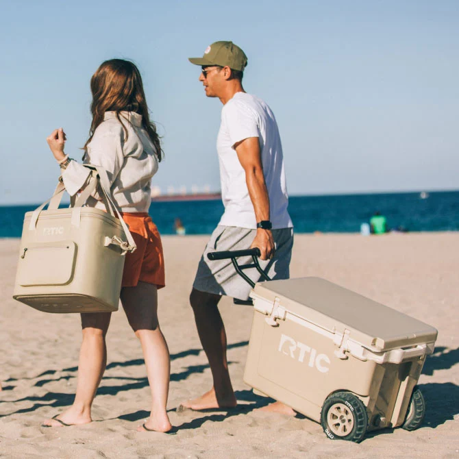 Two people walk on a sandy beach carrying a cooler bag and a wheeled cooler, with the ocean visible in the background.